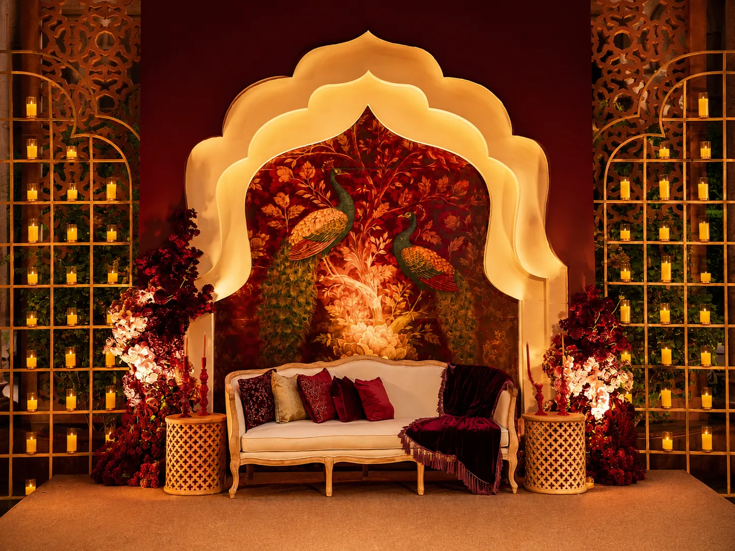 Illuminated Mughal arch backdrop with a hand-painted peacock mural, candlelit gold screens and a cream settee